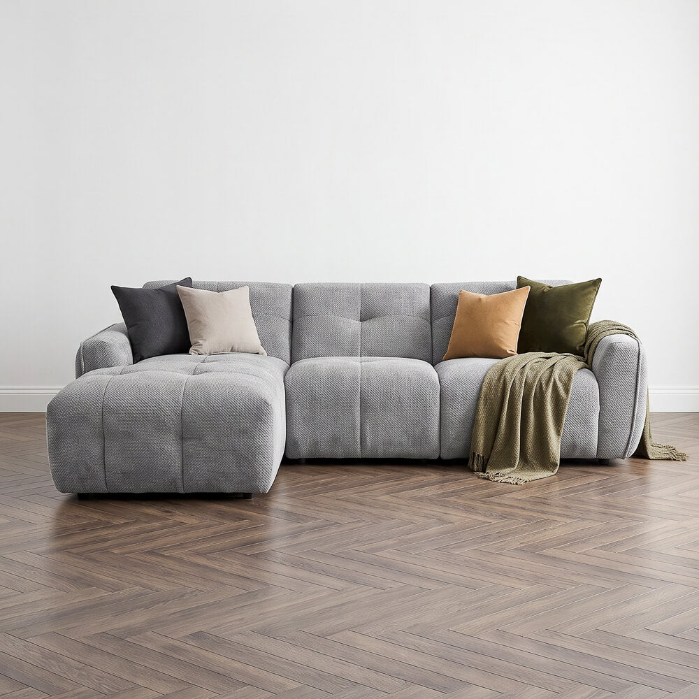 A modern gray tufted sectional sofa with a chaise lounge, styled with multiple accent pillows and a draped throw blanket, set against a minimalist white wall and herringbone wood floor.