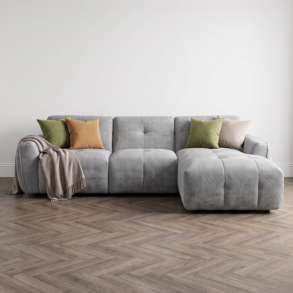 A modern gray tufted sectional sofa with a right-facing chaise lounge, styled with four decorative throw pillows in green, mustard, beige, and gray, and a textured cream throw blanket draped over the left arm. The sofa is set against a plain white wall on a wood herringbone floor.