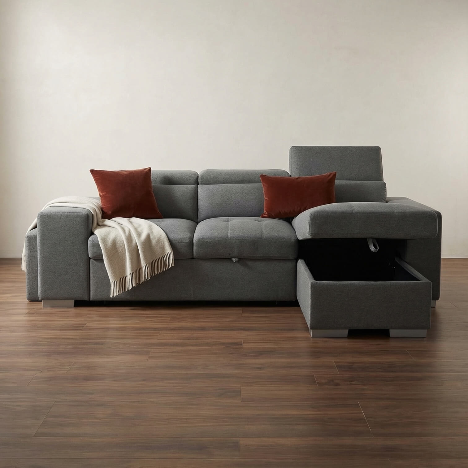 Grey corner sofa bed with red pillows and a blanket on a wooden floor.