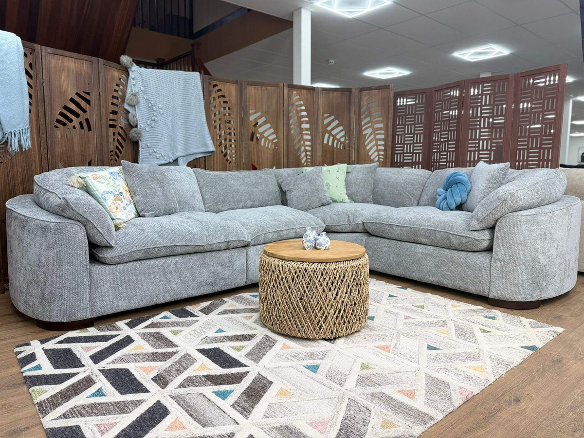 Helena Grey Modular Corner Sofa arranged in an L-shape in a showroom, with patterned throw pillows, a round woven coffee table, and a geometric area rug on a wood floor.