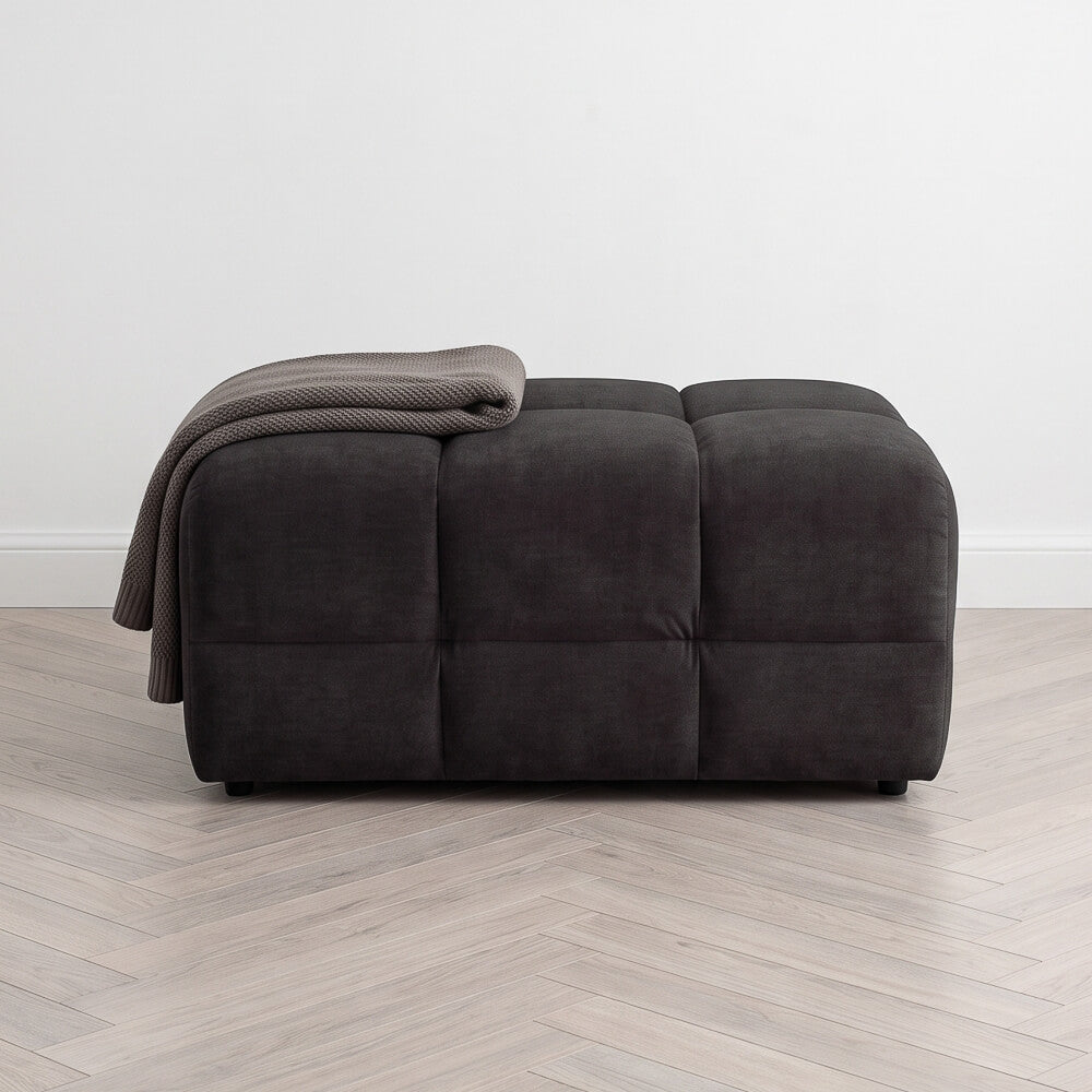 Upholstered dark footstool with tufted panels and folded throw blanket on top