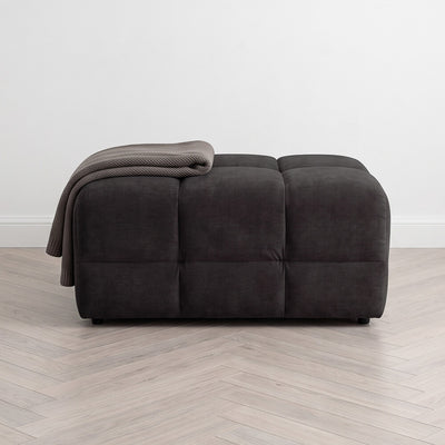 Upholstered dark footstool with tufted panels and folded throw blanket on top